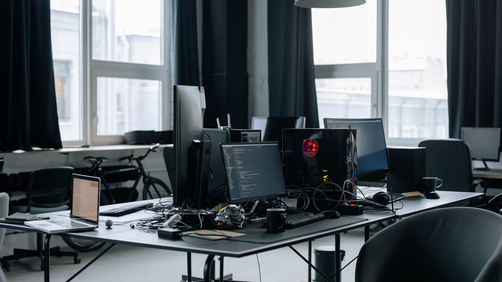 Desktop computers, monitors and cables on desks beside tall office windows.