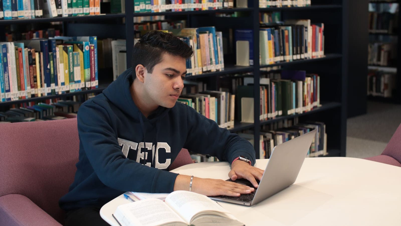 A student uses a laptop beside an open book in a library.
