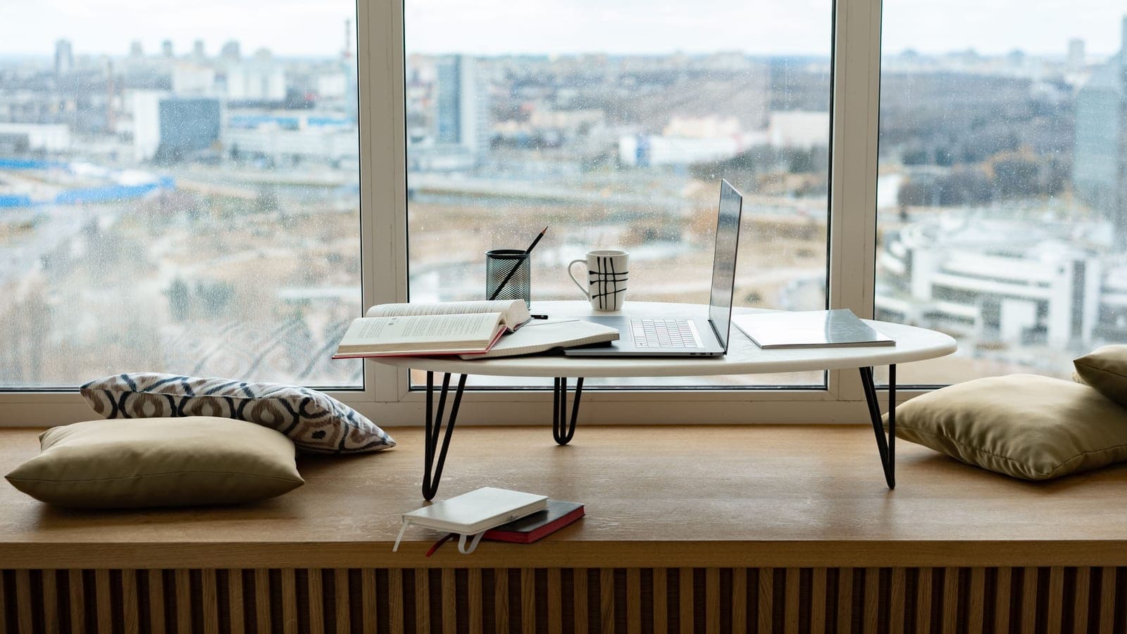 A laptop, open book and mug sit on a table beside a window overlooking a city.